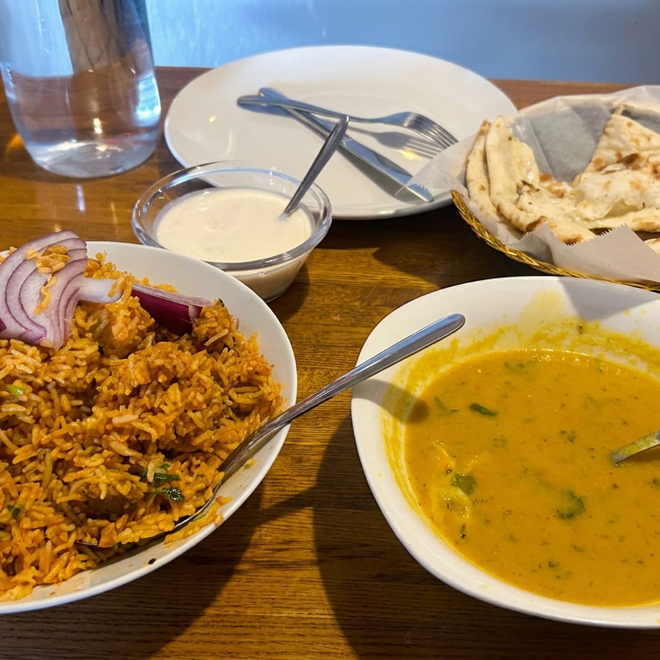 Indian meal with spiced rice, yellow lentil curry, naan bread, yogurt sauce, and sliced red onion on a wooden restaurant table.