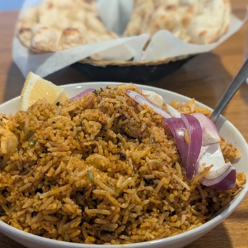 Bowl of spiced biryani rice garnished with red onion and lemon, served with naan and water on a wooden table.