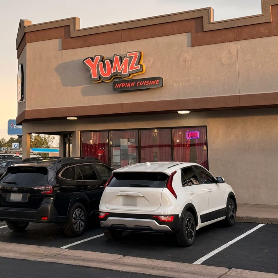 Yumz Indian Cuisine restaurant exterior with illuminated signage, red-curtained windows, entrance, and parked cars.