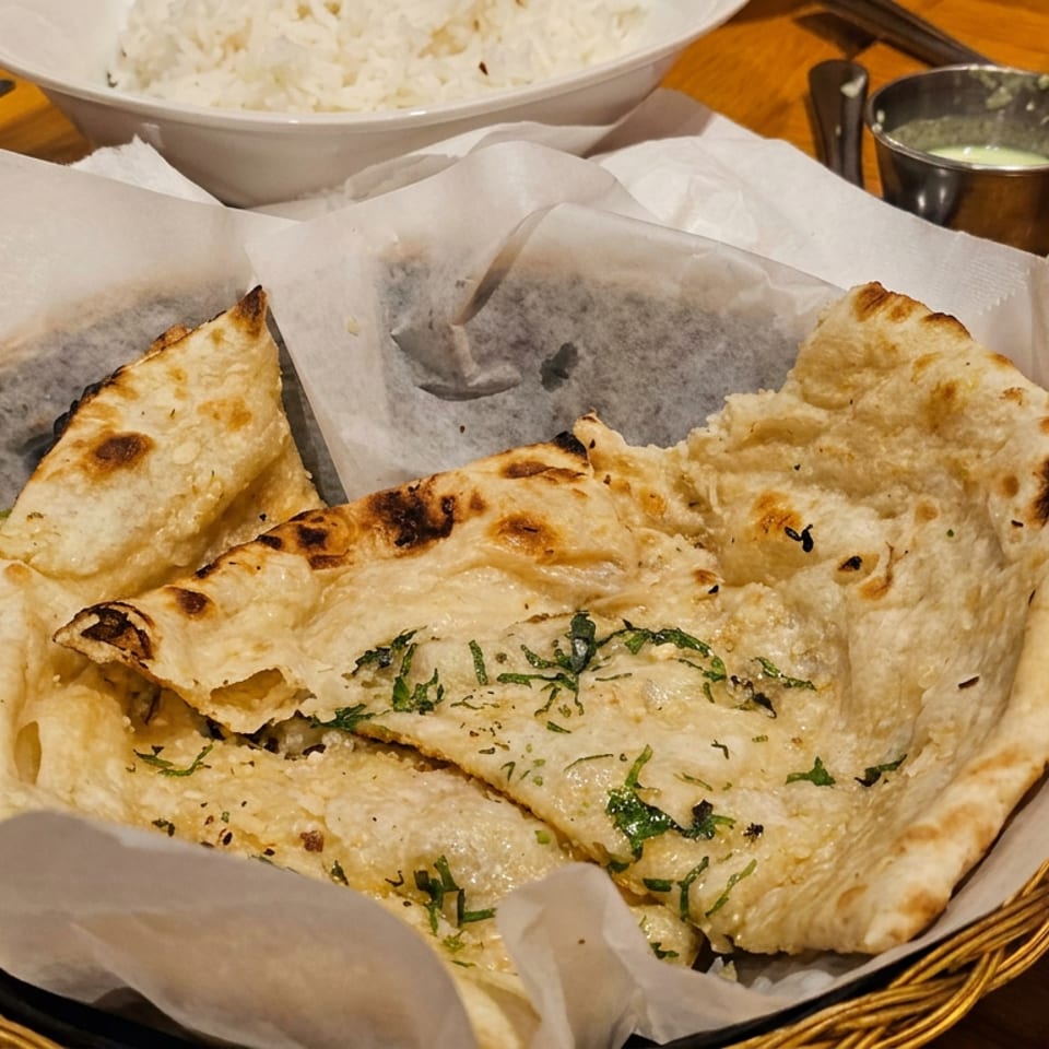 Basket of grilled naan bread garnished with herbs, served beside a bowl of white rice on a wooden restaurant table.
