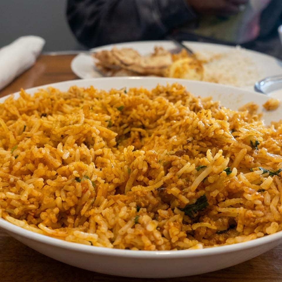 Large bowl of spiced rice with vegetables and herbs, served on a wooden restaurant table with additional dishes and spoon.
