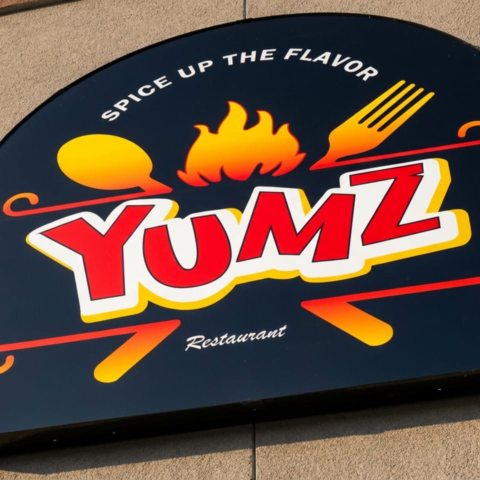 YUMZ Restaurant exterior sign featuring flames, cooking utensils, and the slogan “Spice Up the Flavor.”