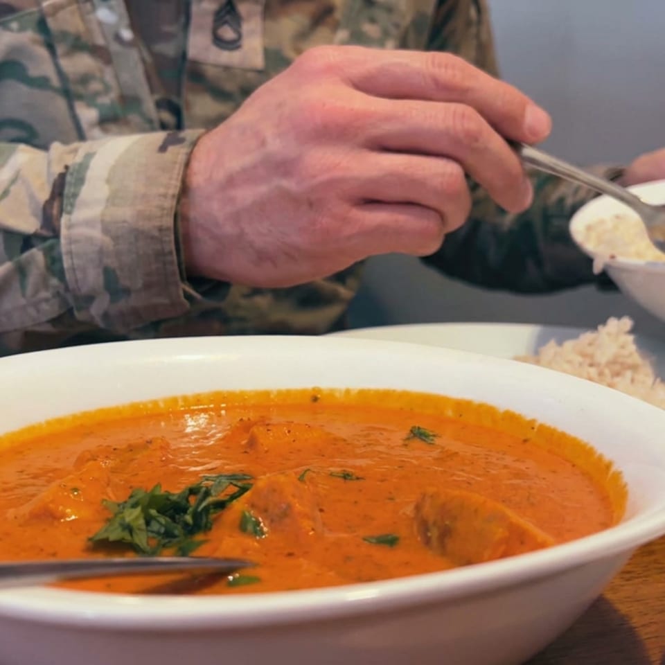Creamy orange curry garnished with herbs sits in a bowl beside rice as a diner in camouflage eats.