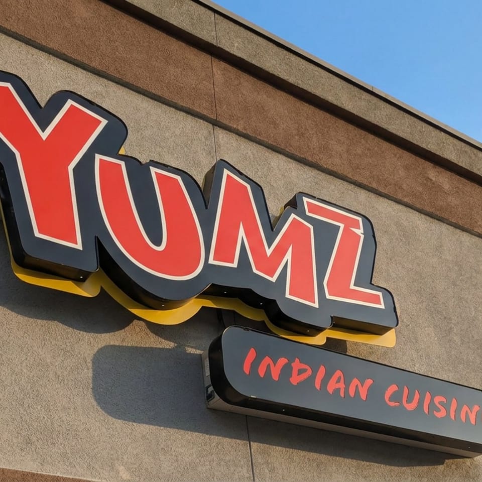 Exterior sign reading YUM Indian Cuisine mounted on a textured restaurant wall beneath a blue sky.