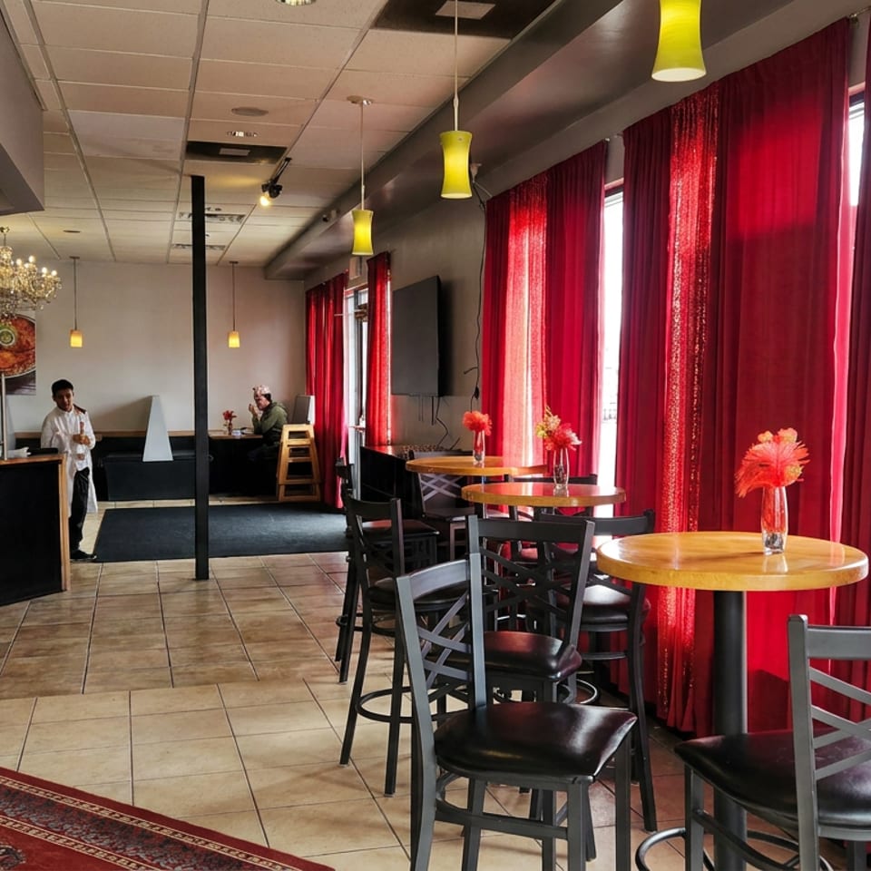 Bright restaurant dining area with red curtains, round wooden tables, black chairs, pendant lights, and a service counter.